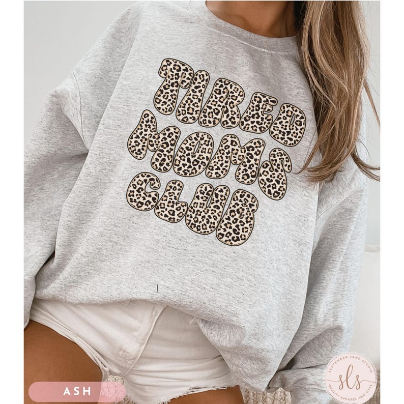 K&ouml;nnte beinhalten: Ein hellgraues Sweatshirt mit dem Text "TIRED MOMS CLUB" in einem Leopardenmuster. Das Sweatshirt hat einen Rundhalsausschnitt und ist aus einem weichen Material gefertigt. Das Wort "ASH" ist unten links aufgedruckt.