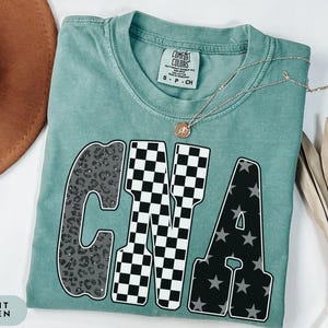 May include: Light green t-shirt with the letters "CNA" in a bold, patterned design. The "C" has a leopard print, the "N" a black and white checkerboard, and the "A" a black background with gray stars. A gold necklace rests on the shirt.