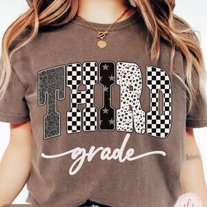 May include: Espresso-colored t-shirt with the words "THIRD grade" in a bold, decorative font. The letters feature various patterns, including leopard print, checkered, and star designs. The word "grade" is written in a cursive script.