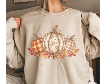May include: A sand-coloured sweatshirt with a seasonal design. The graphic depicts three pumpkins with floral and plaid patterns, surrounded by autumn leaves, acorns, and berries. The word "SAND" is printed at the bottom.