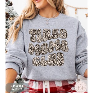 K&ouml;nnte beinhalten: Ein hellgraues Sweatshirt mit dem Text "TIRED MOMS CLUB" in einem Leopardenmuster. Das Sweatshirt hat die Farbe Sportgrau. Am unteren Bildrand ist eine rot-wei&szlig; karierte Hose zu sehen.