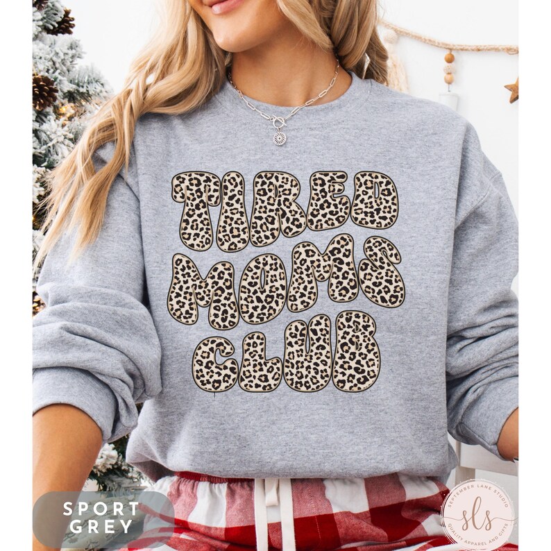 K&ouml;nnte beinhalten: Ein hellgraues Sweatshirt mit dem Text "TIRED MOMS CLUB" in einem Leopardenmuster. Das Sweatshirt hat die Farbe Sportgrau. Am unteren Bildrand ist eine rot-wei&szlig; karierte Hose zu sehen.