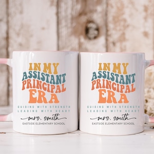 May include: Two white ceramic mugs with pink handles. The mugs feature a colorful graphic that says "In My Assistant Principal Era" with the text "Guiding with Strength Leading with Heart" and "mrs. smith Eastside Elementary School".