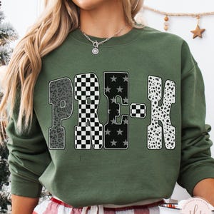 May include: Military green sweatshirt with the word "PRE-K" in a bold, patterned design. The "P" has a leopard print, the "R" has a black and white checkerboard pattern, the "E" has black stars, and the "K" has a dalmatian print.