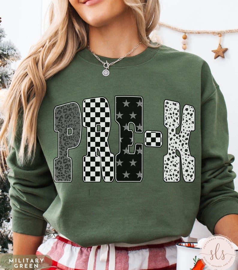 May include: Military green sweatshirt with the word "PRE-K" in a bold, patterned design. The "P" has a leopard print, the "R" has a black and white checkerboard pattern, the "E" has black stars, and the "K" has a dalmatian print.