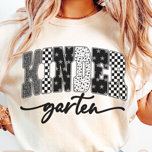 May include: Ivory-colored t-shirt with the word "KINDER" in a bold, multi-patterned design, including leopard print, checkered, and star patterns. The word "garten" is written in a cursive font below the word "KINDER".