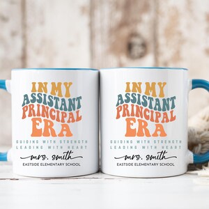 May include: Two white ceramic mugs with blue rims and handles. The mugs feature the text "In My Assistant Principal Era" in a retro font with colorful accents. The text "Guiding with Strength Leading with Heart" is below the main text. The name "mrs. smith" and "Eastside Elementary School" are printed below the text.