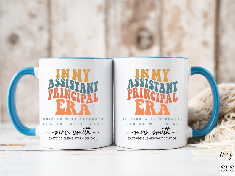 May include: Two white ceramic mugs with blue rims and handles. The mugs feature the text "In My Assistant Principal Era" in a retro font with colorful accents. The text "Guiding with Strength Leading with Heart" is below the main text. The name "mrs. smith" and "Eastside Elementary School" are printed below the text.