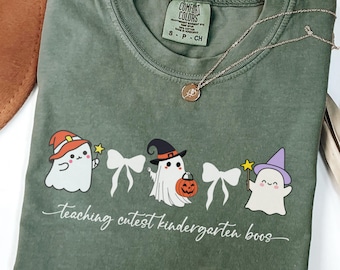 May include: Moss green Comfort Colors t-shirt featuring a Halloween-themed graphic with cartoon ghosts wearing witch hats and holding wands. The text "Teaching cutest kindergarten boos" is printed below the ghosts. A gold necklace is draped across the shirt.