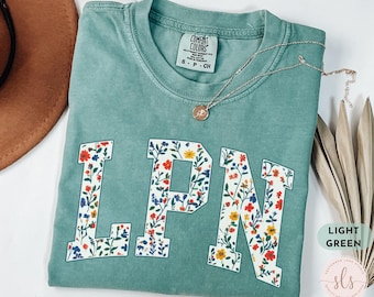 May include: A light green t-shirt with a floral print of the letters LPN. The shirt has a distressed look and is made of a soft, comfortable fabric.