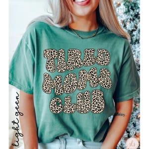 K&ouml;nnte beinhalten: Hellgr&uuml;nes T-Shirt mit dem Text "TIRED MOMS CLUB" in einem Leopardenmuster. Das Shirt hat kurze &Auml;rmel und eine einfarbige Farbe. Die W&ouml;rter sind in einer fetten, abgerundeten Schrift.