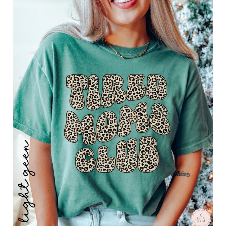 K&ouml;nnte beinhalten: Hellgr&uuml;nes T-Shirt mit dem Text "TIRED MOMS CLUB" in einem Leopardenmuster. Das Shirt hat kurze &Auml;rmel und eine einfarbige Farbe. Die W&ouml;rter sind in einer fetten, abgerundeten Schrift.