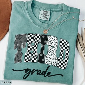 May include: A light green t-shirt with the words "THIRD GRADE" in a bold, patterned design. The word "grade" is written in cursive below the main text. A gold necklace with a pendant rests on the shirt.