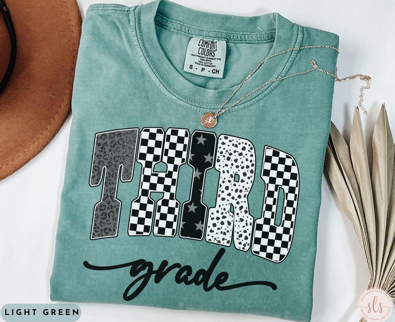 May include: A light green t-shirt with the words "THIRD GRADE" in a bold, patterned design. The word "grade" is written in cursive below the main text. A gold necklace with a pendant rests on the shirt.