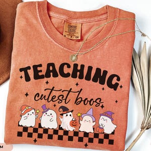 May include: A coral-colored t-shirt with the words "TEACHING cutest boos" in black lettering. Below the text are cartoon ghosts wearing witch hats and holding pumpkins. A gold necklace is draped across the shirt.