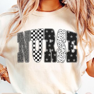 May include: Ivory-colored t-shirt with the word "NURSE" in a bold, multi-patterned design. The letters feature leopard print, checkered, star, and polka dot patterns. The word "IVORY" is printed at the bottom of the shirt.