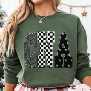 May include: Olive green sweatshirt with the letters "CNA" in a bold, decorative font. The "C" has a leopard print design, the "N" is checkered, and the "A" features a star pattern. The text "MILITARY GREEN" is at the bottom left.
