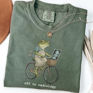 May include: Moss green t-shirt featuring a frog riding a bicycle and holding an X-ray. The frog wears an apron and glasses. The text "off to radiology" is printed at the bottom. A gold necklace is draped across the shirt.