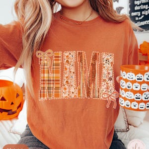 May include: An orange-colored sweatshirt with the word "MAMA" in a decorative design. The letters feature plaid, floral, and textured patterns. A matching orange mug with jack-o'-lantern faces is held in the hand. The sweatshirt is a fall-themed item.