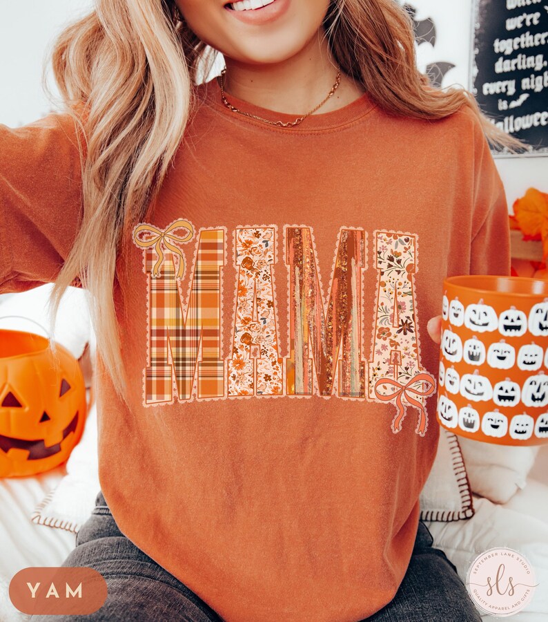 May include: An orange-colored sweatshirt with the word "MAMA" in a decorative design. The letters feature plaid, floral, and textured patterns. A matching orange mug with jack-o'-lantern faces is held in the hand. The sweatshirt is a fall-themed item.