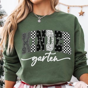 May include: A military green sweatshirt with the word "KINDERGARTEN" in a bold, patterned design. The word "kindergarten" is in a white script font. The sweatshirt is a comfortable, casual style.