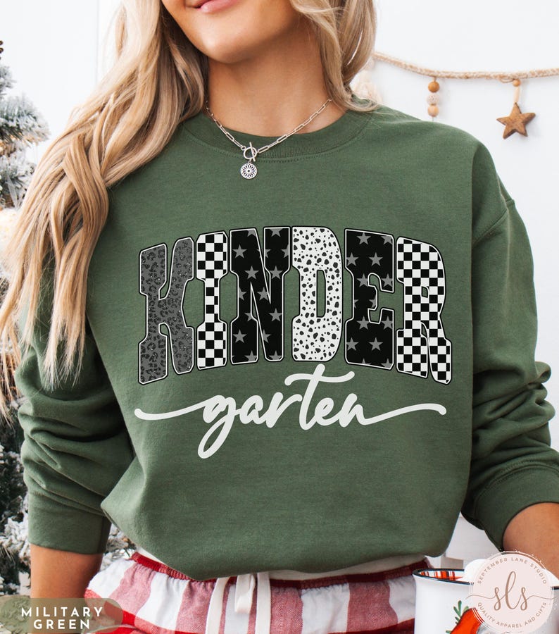 May include: A military green sweatshirt with the word "KINDERGARTEN" in a bold, patterned design. The word "kindergarten" is in a white script font. The sweatshirt is a comfortable, casual style.
