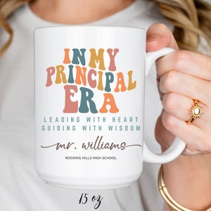 以下が含まれることがあります： カラフルなレトロなフォントで「IN MY PRINCIPAL ERA」という文字が書かれた白いセラミックマグカップ。メインテキストの下には「LEADING WITH HEART GUIDING WITH WISDOM」というテキストがあります。「mr. williams」というテキストは、テキストの下に筆記体で書かれています。「ROCKING HILLS HIGH SCHOOL」というテキストは、筆記体テキストの下にあります。マグカップは15オンスです。