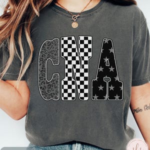 May include: Dark grey t-shirt with the letters "CNA" in a bold, outlined font. The "C" has a leopard print design, the "N" is checkered, and the "A" has a star pattern. The word "PEPPER" is at the bottom.