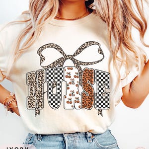 May include: Ivory-colored t-shirt with the word "NURSE" in a bold, decorative font. The letters feature leopard print, checkered patterns, and cherry designs. A large bow with a leopard print pattern is above the word.