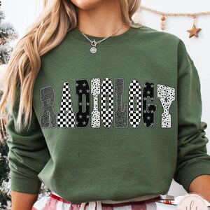 K&ouml;nnte beinhalten: Milit&auml;rgr&uuml;nes Sweatshirt mit dem Wort "RADIOLOGY" in einem auff&auml;lligen, gemusterten Design. Die Buchstaben zeigen eine Mischung aus Leopardenmuster, Schachbrettmuster und Sternenmustern. Das Sweatshirt ist ein Rundhals-Stil.