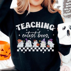 May include: Black sweatshirt with the text "TEACHING cutest boos" in white. Below the text are six cartoon ghosts wearing hats and holding pumpkins. A white and black checkered pattern is below the ghosts. A pumpkin and a ghost mug are also visible.