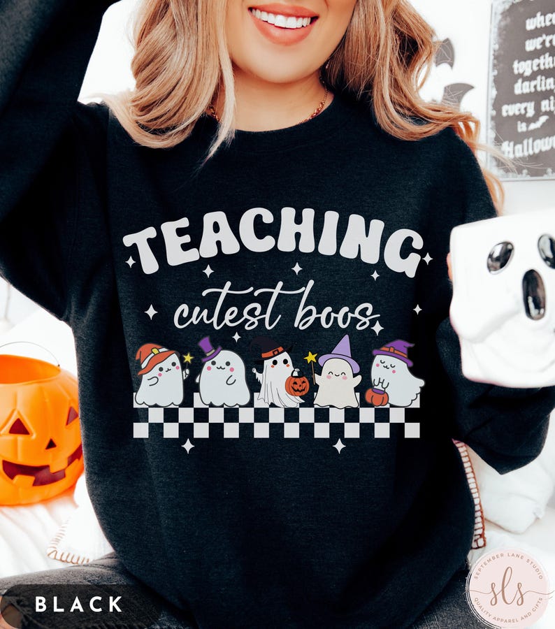 May include: Black sweatshirt with the text "TEACHING cutest boos" in white. Below the text are six cartoon ghosts wearing hats and holding pumpkins. A white and black checkered pattern is below the ghosts. A pumpkin and a ghost mug are also visible.