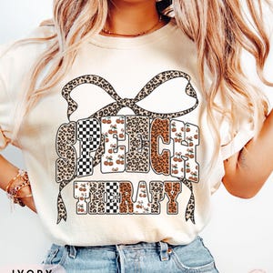 Peut inclure: T-shirt ivoire avec les mots "SPEECH THERAPY" dans un mélange d'imprimé léopard, de damier et de motifs de cerises. Un nœud léopard est au-dessus du texte. Le mot "IVORY" est en bas.