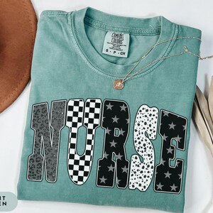 May include: Light green t-shirt with the word "NURSE" in a bold, block letter design. The letters feature various patterns, including leopard print, checkered, stars, and polka dots. A gold necklace with a pendant rests on the shirt.