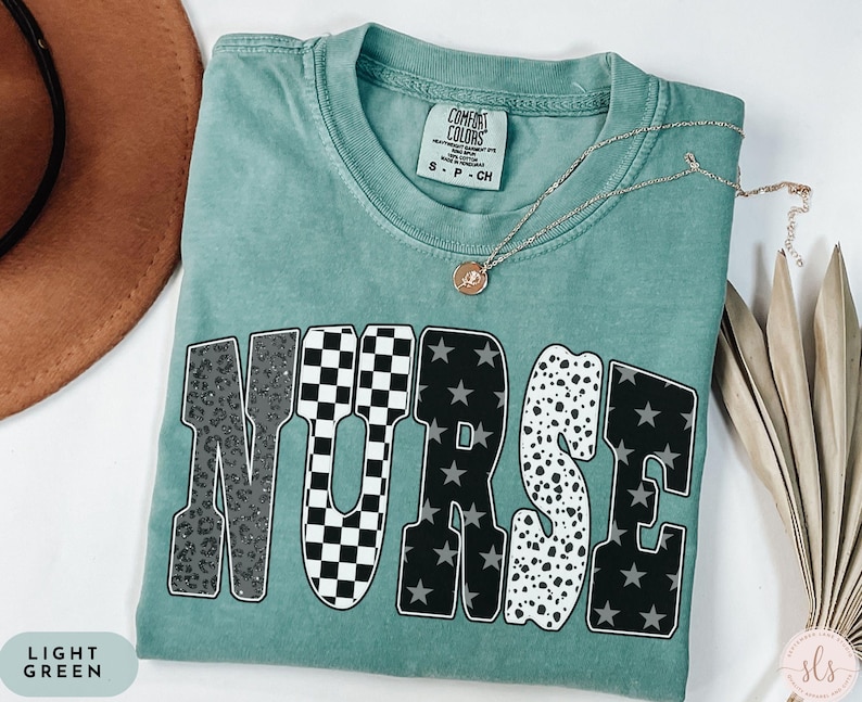 May include: Light green t-shirt with the word "NURSE" in a bold, block letter design. The letters feature various patterns, including leopard print, checkered, stars, and polka dots. A gold necklace with a pendant rests on the shirt.