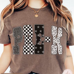 K&ouml;nnte beinhalten: Espressofarbenes T-Shirt mit dem Wort "PRE-K" in einem auff&auml;lligen, gemusterten Design. Die Buchstaben zeigen Leopardenmuster, ein schwarz-wei&szlig;es Schachbrettmuster, Sterne und Dalmatinerflecken. Eine goldene Halskette ist sichtbar.