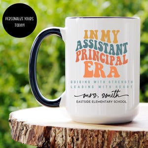 May include: White ceramic coffee mug with a black handle. The mug has a colorful graphic that says "In My Assistant Principal Era" with the text "Guiding with Strength Leading with Heart" and "mrs. smith Eastside Elementary School".