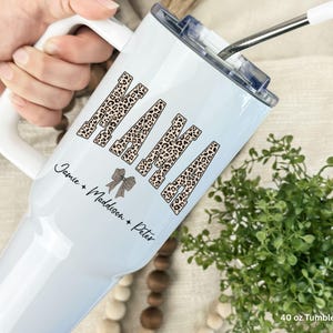 May include: A white 40 oz tumbler with a handle and a metal straw. The tumbler features the word NANA in a leopard print pattern, followed by a small bow icon and the names Jamie, Maddison, and Peter printed in black script lettering.