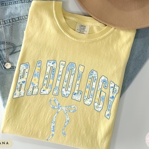 May include: A yellow t-shirt with the word "RADIOLOGY" in blue letters with a floral pattern. A blue bow is below the word. The shirt is folded on top of blue jeans.