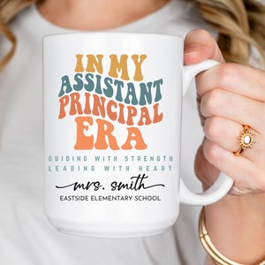 May include: White ceramic coffee mug with the text "IN MY ASSISTANT PRINCIPAL ERA GUIDING WITH STRENGTH LEADING WITH HEART mrs. smith EASTSIDE ELEMENTARY SCHOOL" in colorful retro font.