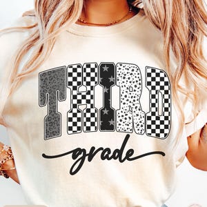 May include: Ivory-colored t-shirt with the words "THIRD grade" in a bold, decorative font. The word "THIRD" features a mix of patterns, including leopard print, checkered, and stars. The word "grade" is written in a cursive script.