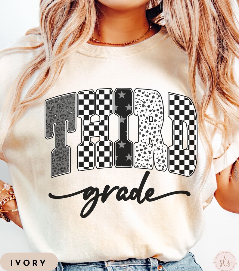 May include: Ivory-colored t-shirt with the words "THIRD grade" in a bold, decorative font. The word "THIRD" features a mix of patterns, including leopard print, checkered, and stars. The word "grade" is written in a cursive script.