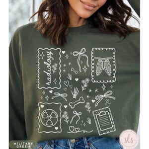 Puede incluir: Sudadera verde militar con un dise&ntilde;o gr&aacute;fico blanco que presenta la palabra "radiolog&iacute;a", huesos, corazones, lazos, flores y una caja tor&aacute;cica. La sudadera tiene cuello redondo y mangas largas.
