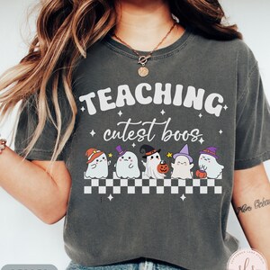 May include: Dark gray t-shirt with the words "TEACHING cutest boos" in white. Below the text are five cartoon ghosts wearing different Halloween hats. The shirt has a checkerboard pattern at the bottom and the word "PEPPER" at the bottom left.