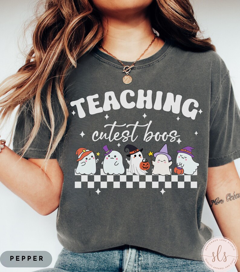 May include: Dark gray t-shirt with the words "TEACHING cutest boos" in white. Below the text are five cartoon ghosts wearing different Halloween hats. The shirt has a checkerboard pattern at the bottom and the word "PEPPER" at the bottom left.