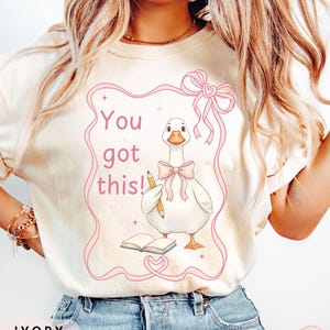 May include: Ivory-colored t-shirt featuring a cartoon goose with a pink bow, holding a pencil, and standing near an open book. The text "You got this!" is in pink script within a pink ribbon frame. The shirt is a casual style.