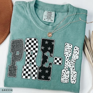 May include: Light green t-shirt with the word "PRE-K" in a mix of patterns: leopard print, checkered, stars, and dalmatian spots. A gold necklace with a pendant rests on the shirt. The shirt is labeled "Comfort Colors".