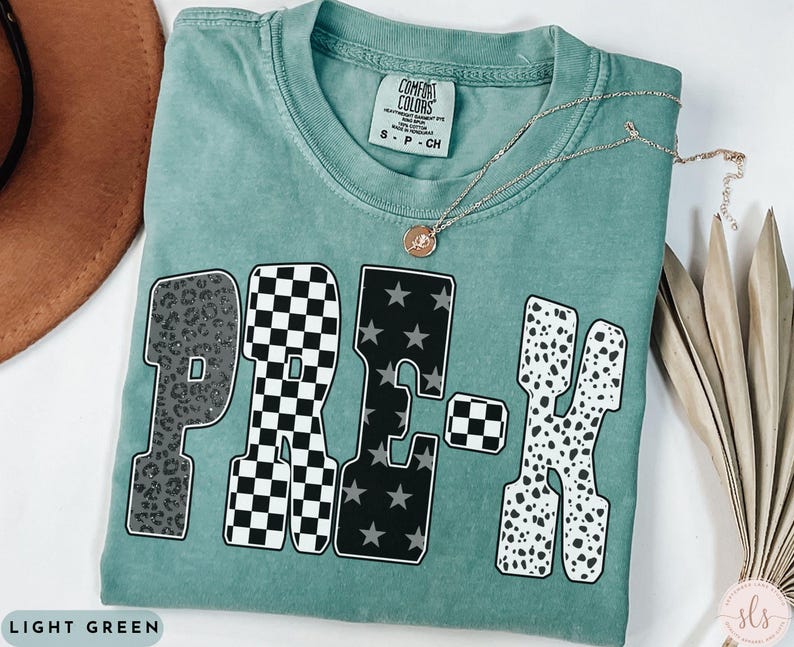 May include: Light green t-shirt with the word "PRE-K" in a mix of patterns: leopard print, checkered, stars, and dalmatian spots. A gold necklace with a pendant rests on the shirt. The shirt is labeled "Comfort Colors".