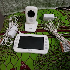 May include: A white Axvue baby monitor set including a camera unit with a black lens, a rectangular handheld monitor with a screen and control buttons, and two white power adapters with attached cords for charging the electronic devices.