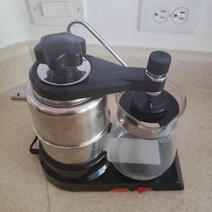 May include: A countertop coffee maker featuring a cylindrical stainless steel water reservoir with a large black knob on top, connected to a glass carafe by a black plastic arm. The base is black with orange and red power switches.